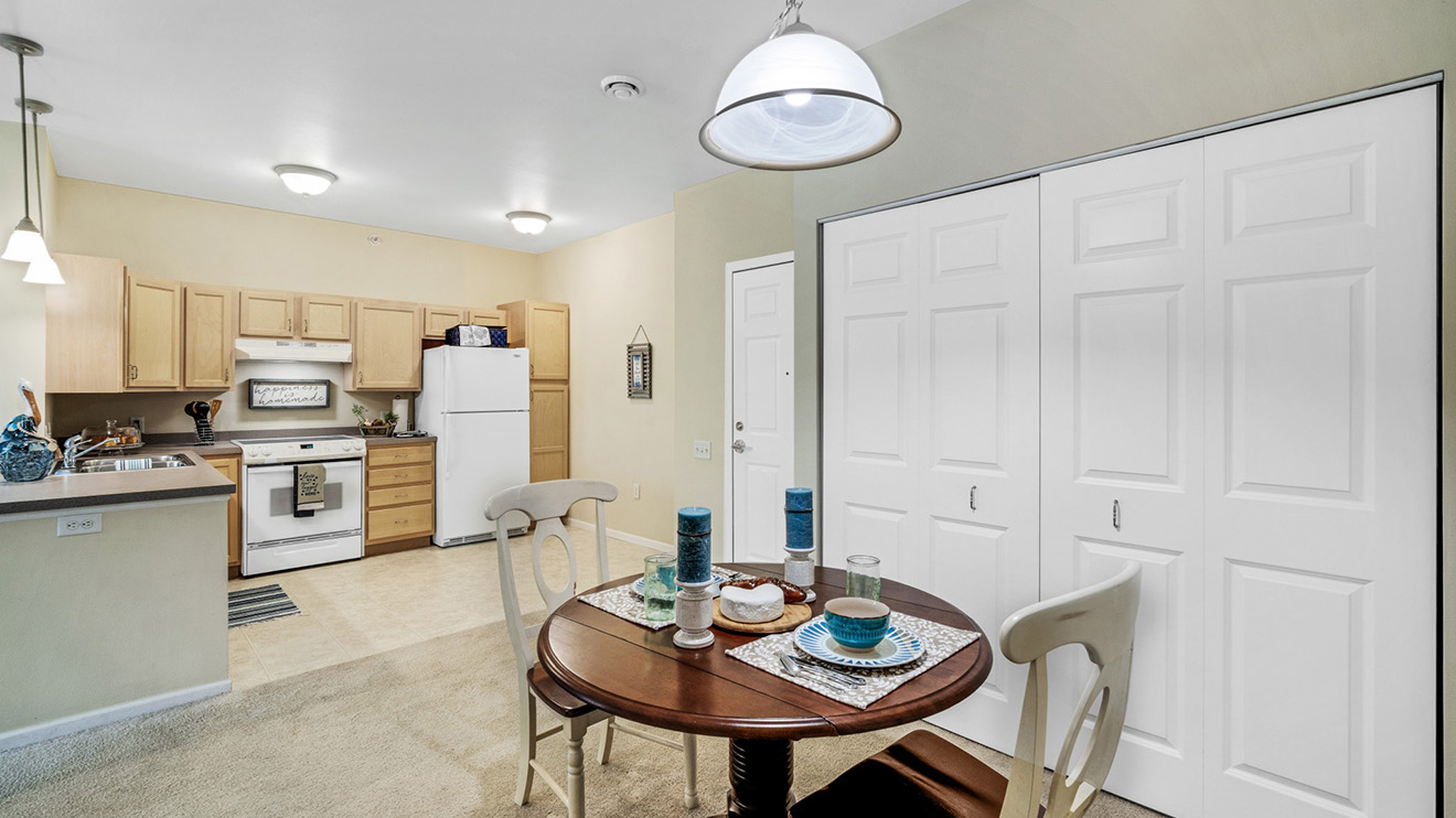 Holiday Village at the Falls senior apartment with kitchen and white appliances, closet and a dinette set under overhead light.